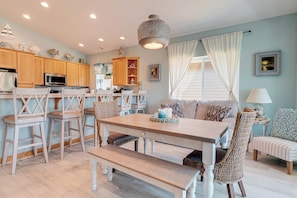 A dining area with a wooden table and chairs, a hanging light fixture, and a view of the kitchen in the background. - Friends & Family Seaside Retreat, Walk to Beach! (Gold Beach)