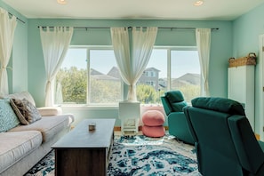 A living room with a sofa, a chair, a coffee table, and a view of houses through large windows. - Friends & Family Seaside Retreat, Walk to Beach! (Gold Beach)