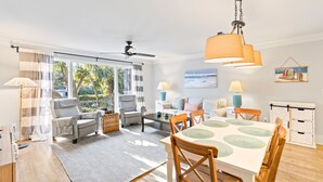 A modern living room with a dining area, large windows, and a ceiling fan. - Lagoon Villa 43 - Walk to Ocean! Prime Location! (Isle of Palms)