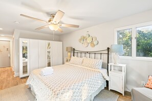 A bedroom with a bed, a ceiling fan, a nightstand with a lamp, a window, and a door leading to a bathroom.