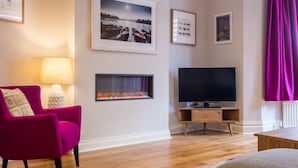 Ein Wohnzimmer mit Kamin, Flachbildfernseher, einem rosa Sessel und gerahmten Bildern an der Wand.