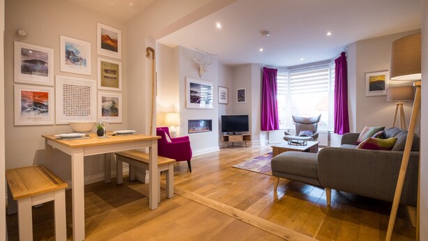 Ein modernes Wohnzimmer mit Holzboden, einem Essbereich mit Tisch und Stühlen, einem grauen Sofa und einem Fernseher.