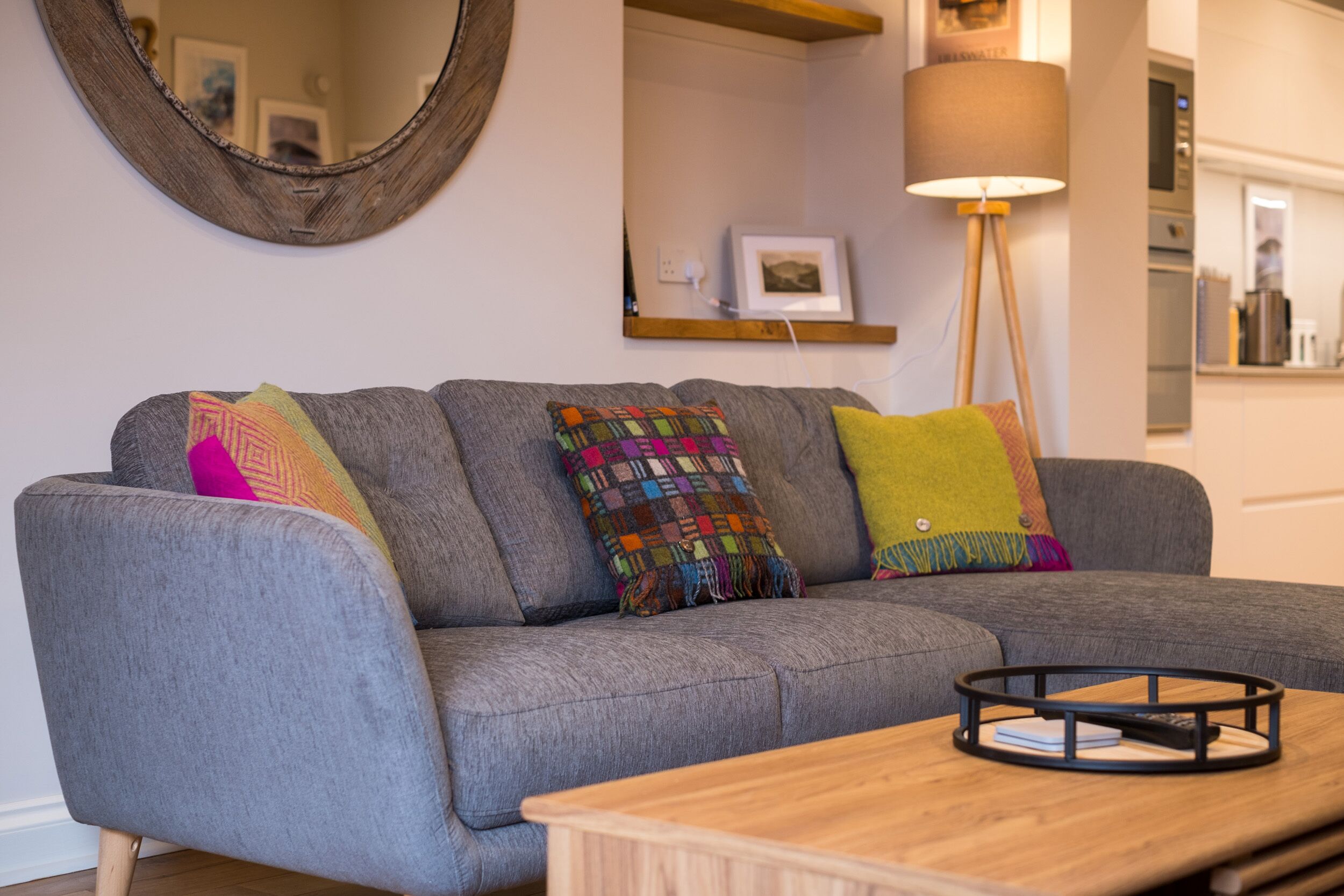 A grey sofa with colorful cushions, a wooden coffee table, a round mirror, and a floor lamp.