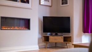 Ein modernes Wohnzimmer mit Fernseher auf einem Holzständer, einem Kamin mit brennenden Kerzen und gerahmten Bildern an der Wand.