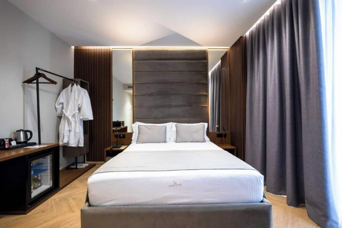 A modern hotel room with a large bed, a wardrobe, a desk with a kettle, and a view of the city.