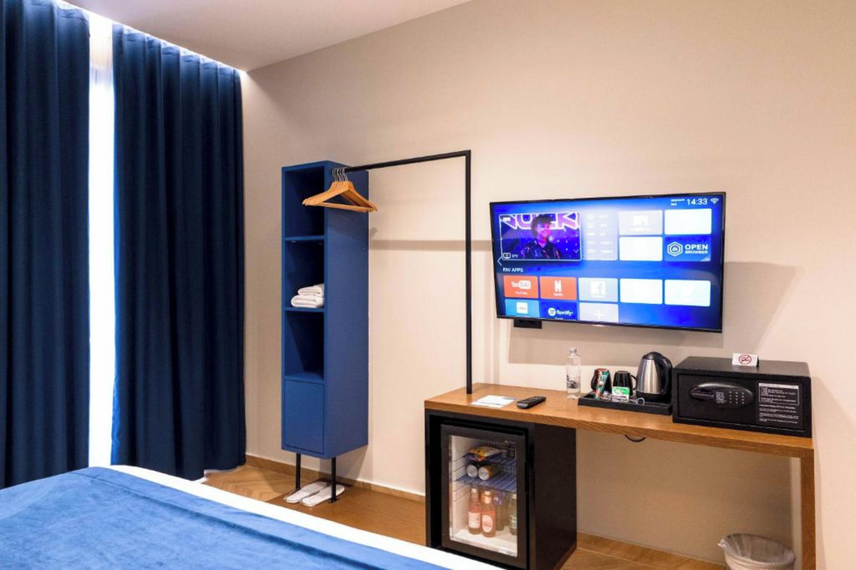 A hotel room with a bed, a desk with a computer monitor, a small fridge, a microwave, and a hanging coat rack.