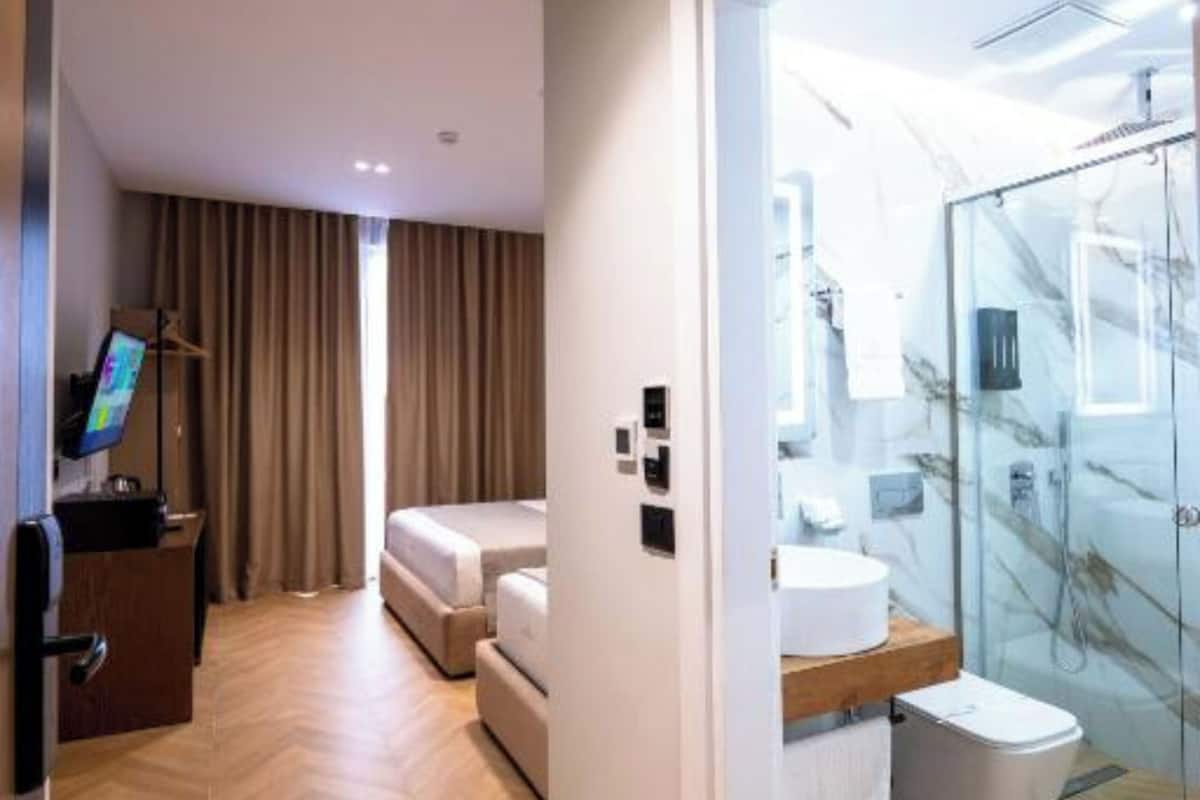 A hotel room with a wooden floor, a bed, a desk with a computer, a TV, and a bathroom with a glass shower.
