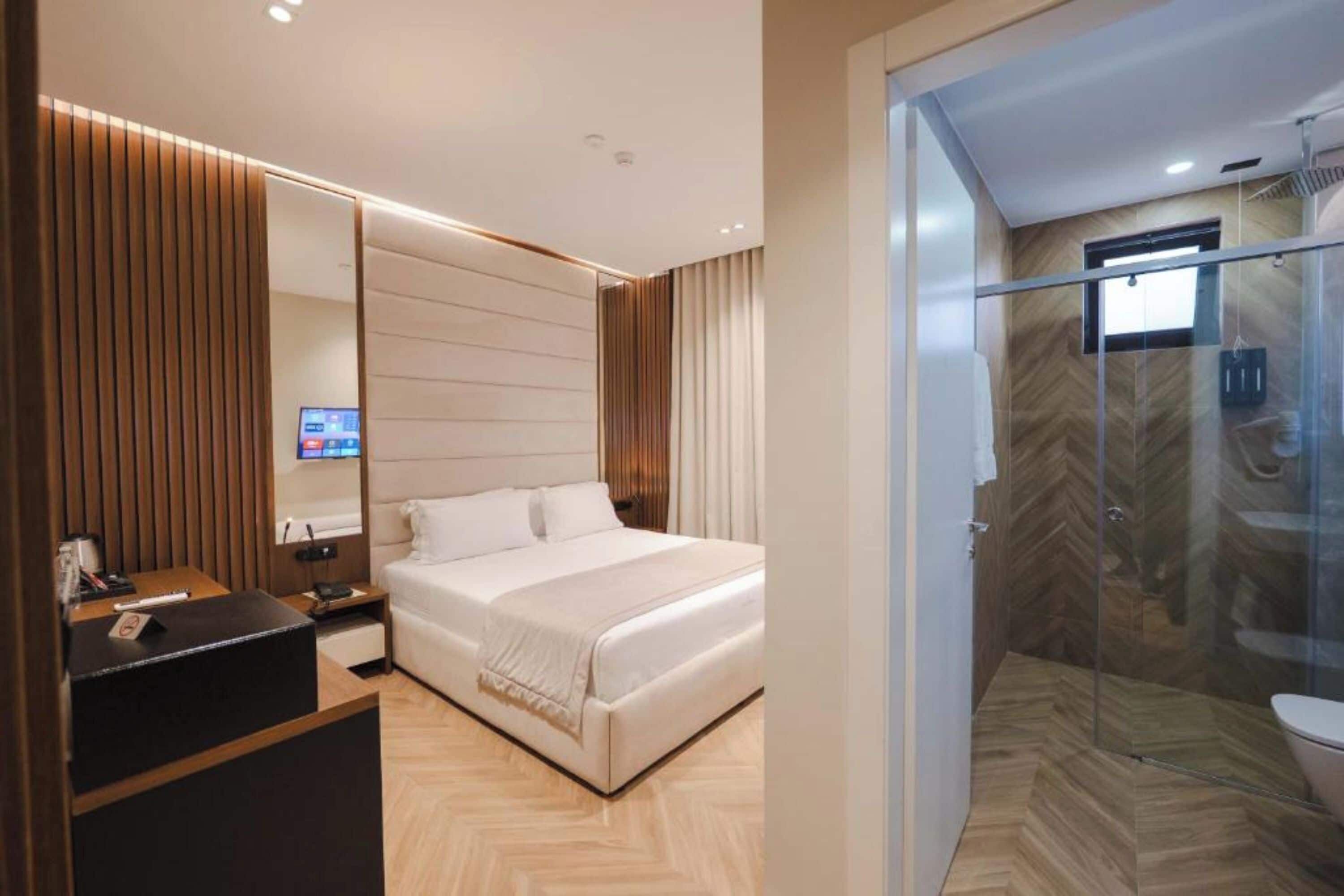 A modern hotel room with a large bed, a desk with a computer, a glass-enclosed shower, and a wooden floor.