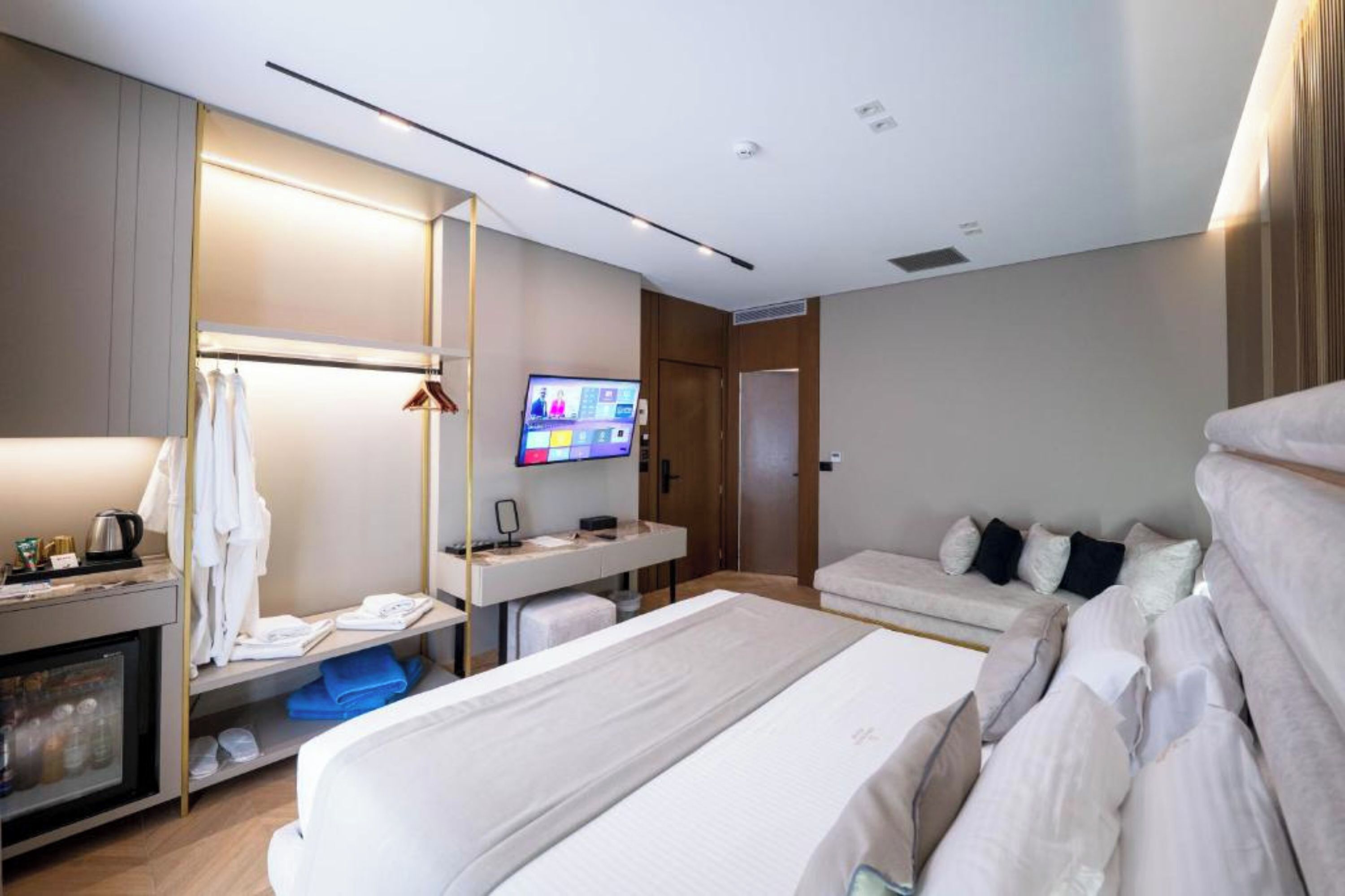 A modern hotel room with a bed, a TV, a desk with a computer, a wardrobe, and a minibar.