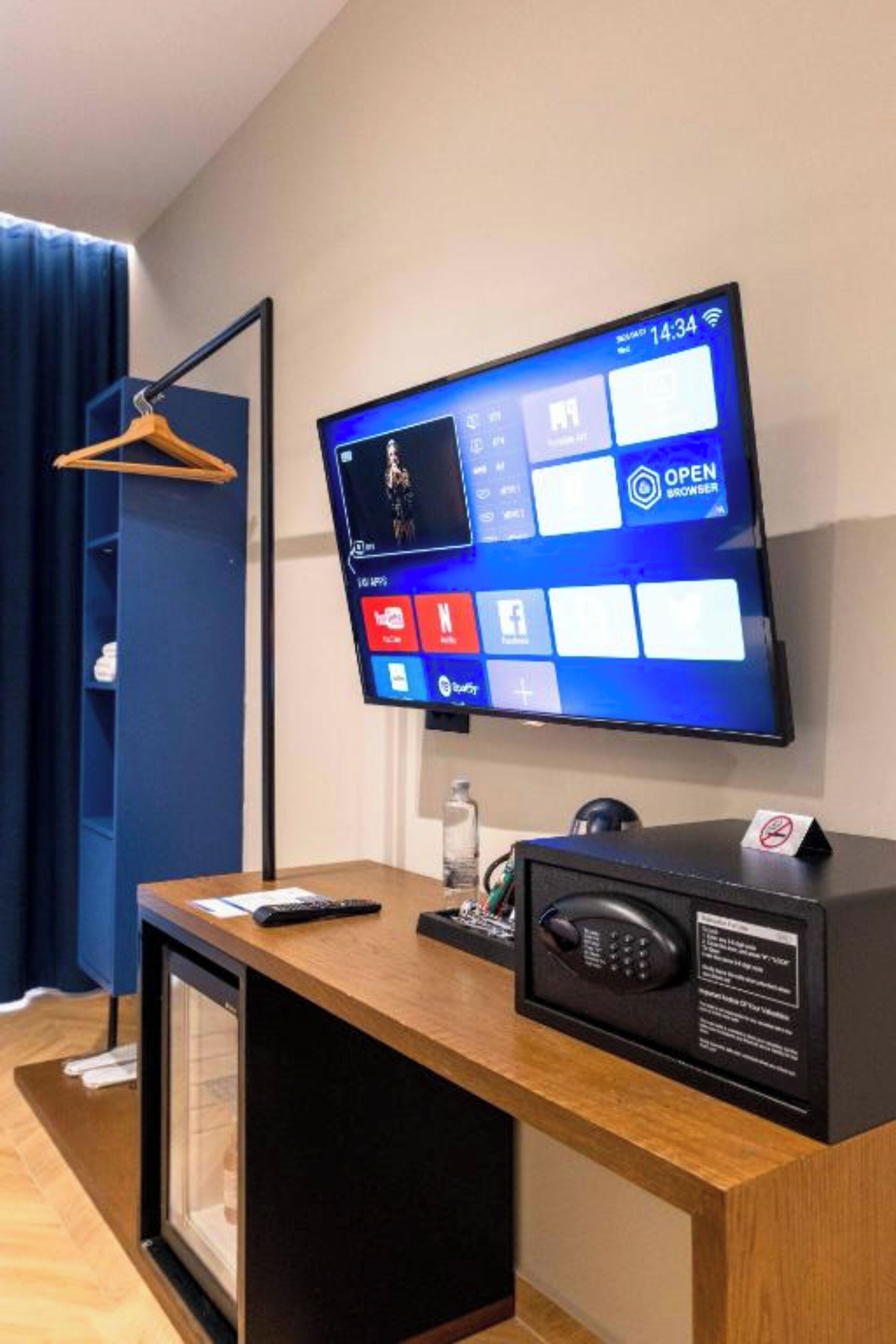 Hotelkamer met een wandtelevisie waarop de Windows interface te zien is, een bureau met een afstandsbediening, een telefoon en een klein tafeltje met een glazen vaas.