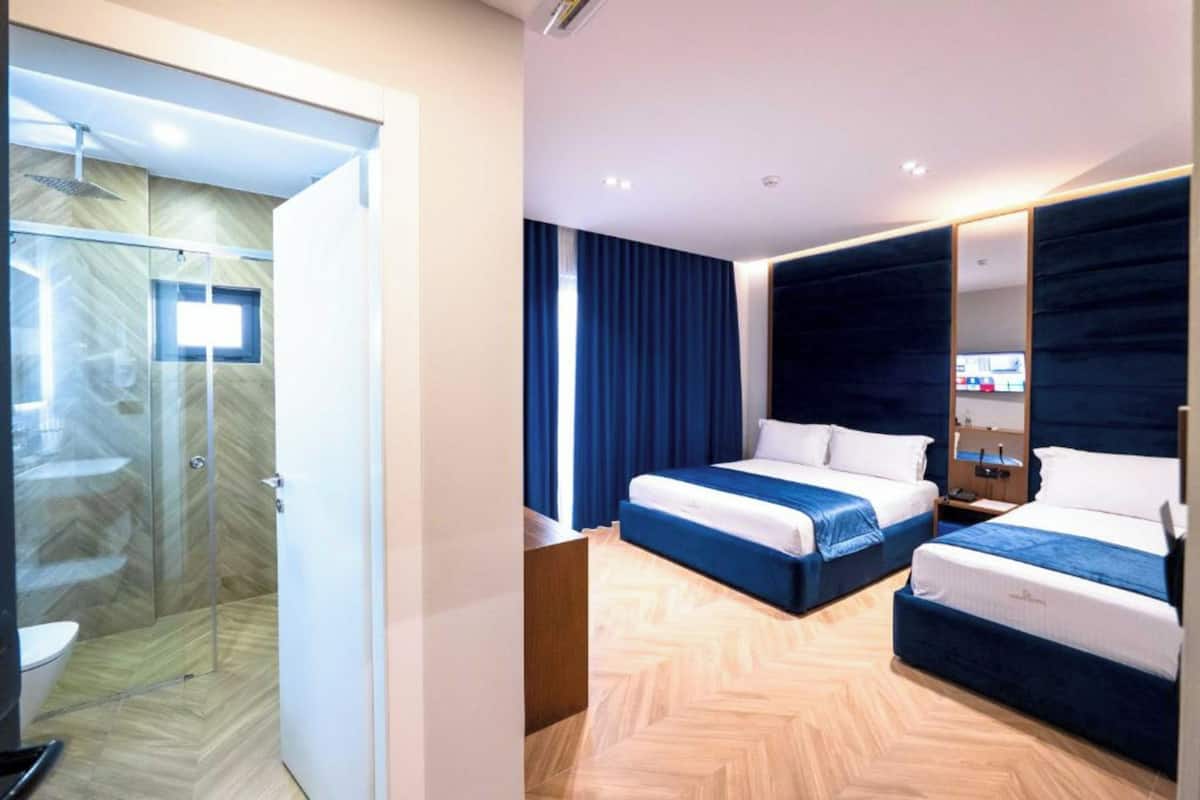 A hotel room with a glass shower enclosure, two beds with blue bedding, a large mirror, and a herringbone-patterned floor.