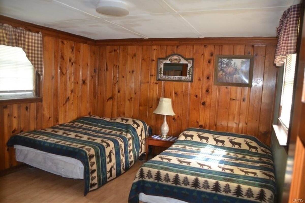 Ein Zimmer mit holzverkleideten Wänden, zwei Betten mit Rentier-Muster-Bettwäsche, ein kleiner Tisch mit einer Lampe, ein gerahmtes Willkommensschild und ein Bild an der Wand.