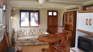 Un salon avec un canapé à motifs floraux, une table à manger en bois avec des chaises, un ventilateur de plafond, un réfrigérateur et un buffet en bois.