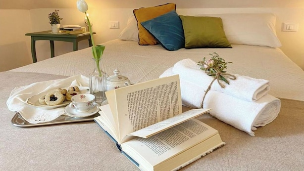 A neatly made bed with pillows, a book, a tray with cookies, a vase with flowers, and a small table with a lamp and a plant. - Juniper House via Mazzini (Torino)