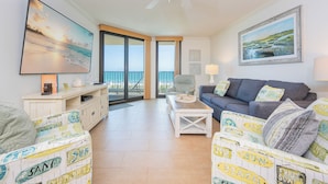Un salon spacieux avec un balcon, un ventilateur de plafond et un grand tableau représentant une scène de plage.
