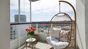 A balcony with a hanging chair, a small table with a vase of flowers, and a view of the cityscape.