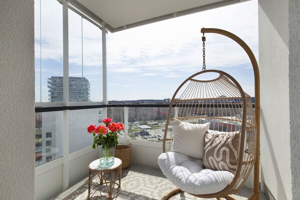A balcony with a hanging chair, a small table with a vase of flowers, and a view of the cityscape.