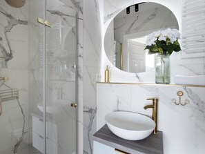 A bathroom with a glass-enclosed shower, a marble wall, a white sink with a gold faucet, and a mirror with a vase of flowers.