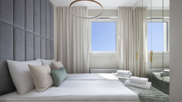 A modern bedroom with a large bed, a view of the sky through a window, and a mirror reflecting the room.