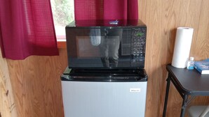 Ein kompakter Kühlschrank mit einer Mikrowelle darüber, ein kleiner Tisch mit Taschentuchhalterung und einer Flasche, sowie ein Fenster mit roten Vorhängen.
