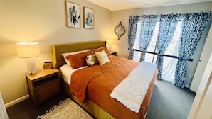 A neatly made bed with a brown quilt, a white throw, and a small animal pillow. There are two bedside lamps, a small nightstand, and a framed picture above the bed. The room has a window with patterned curtains and a view of the outside.
