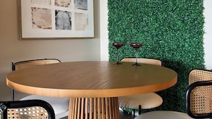 A round wooden table with a woven base, surrounded by four chairs with woven backs. A framed abstract art piece is on the wall, and a green wall with a neon sign is behind the table.