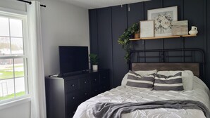 A bedroom with a bed, a dresser, a TV, a shelf with decorations, and a window.