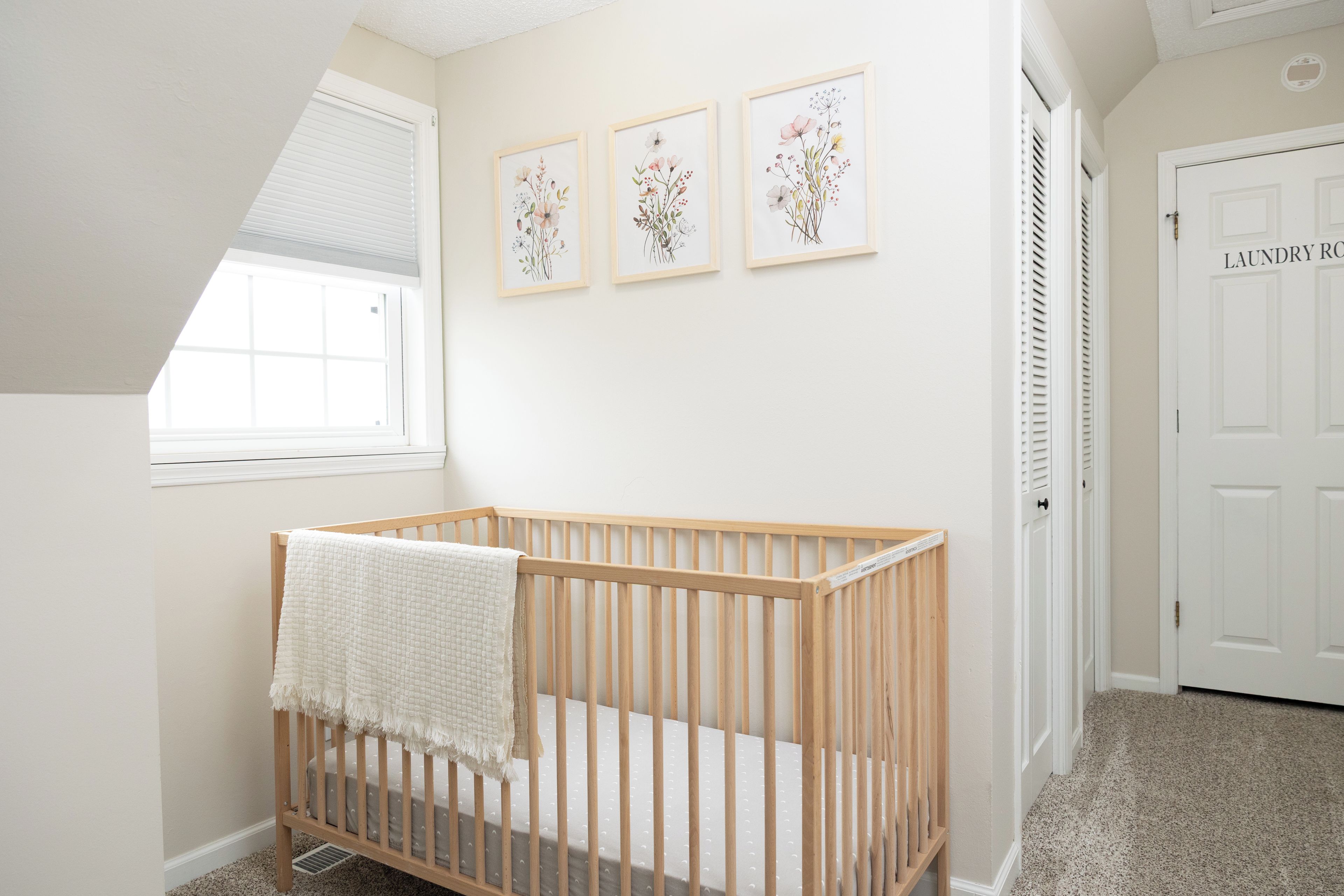 A baby crib with a white blanket, a window, and three framed floral artworks on the wall.