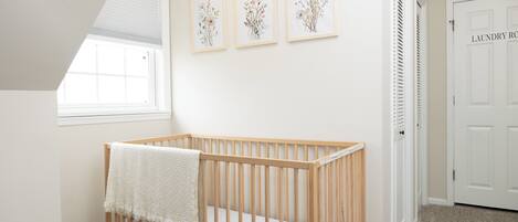 A baby crib with a white blanket, a window, and three framed floral artworks on the wall.