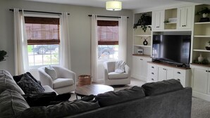 A living room with a grey sofa, white armchairs, a coffee table, and a TV stand with a flat-screen TV. There are white cabinets and shelves with decorative items.