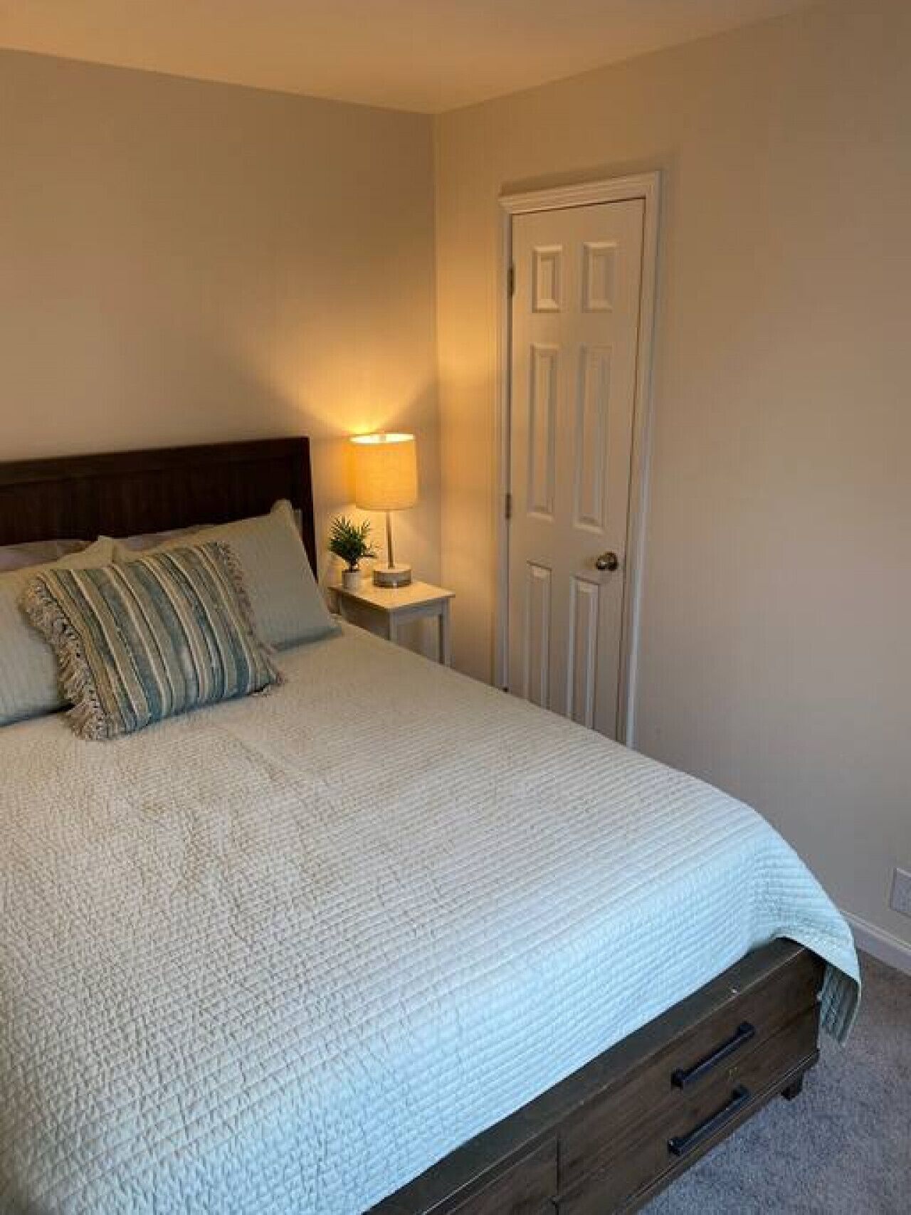 Een net opgemaakt bed met een witte quilt, een bedlampje en een nachtkastje van hout.