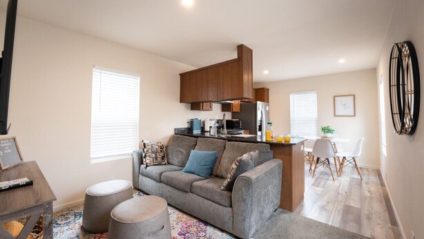 A modern living room with a grey sofa, ottomans, and a colorful rug. A kitchen area is visible with wooden cabinets and a refrigerator.