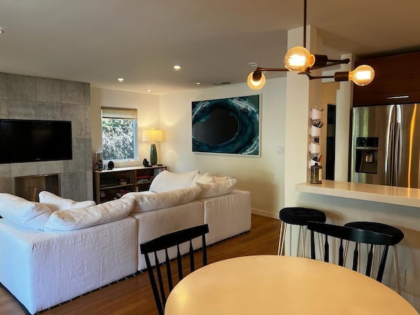 A modern living room with a sectional sofa, a flat-screen TV, a dining area with a round table, and a kitchen with a stainless steel refrigerator.