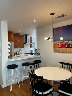 A modern kitchen with wooden cabinets, a white countertop, and a built-in stove. There is a dining area with a round table and four bar stools.