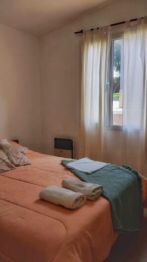 Un dormitorio con cama, almohadas, una mesita de noche y un ventanal con cortinas.