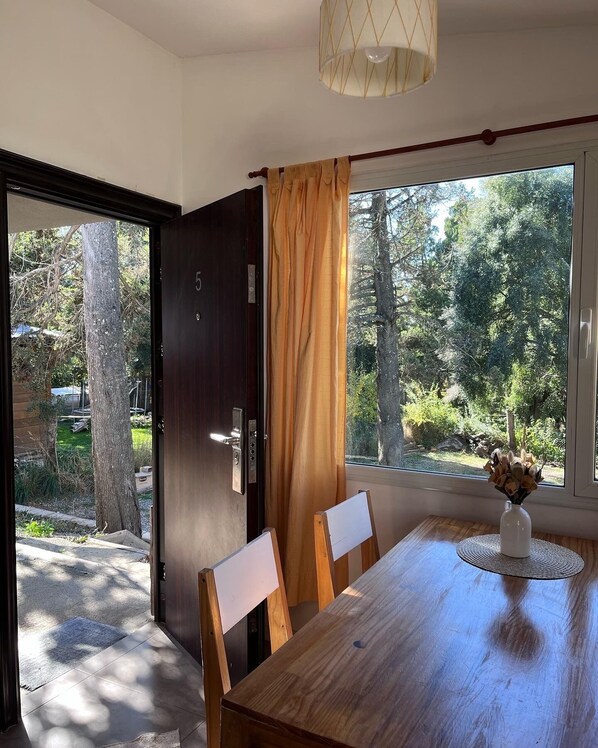 Un comedor con una mesa de madera, sillas, un florero con flores y vistas a un jardín a través de una ventana con cortinas.