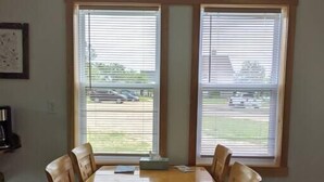 A dining area with a wooden table and chairs, large windows with blinds, and a view of a building and parking lot outside. - Aurora Cottage At The Spruces (Ingonish)
