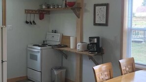 A compact kitchen with a stove, sink, and a small trash bin. There is a wooden table and chairs, and a window with blinds. - Aurora Cottage At The Spruces (Ingonish)