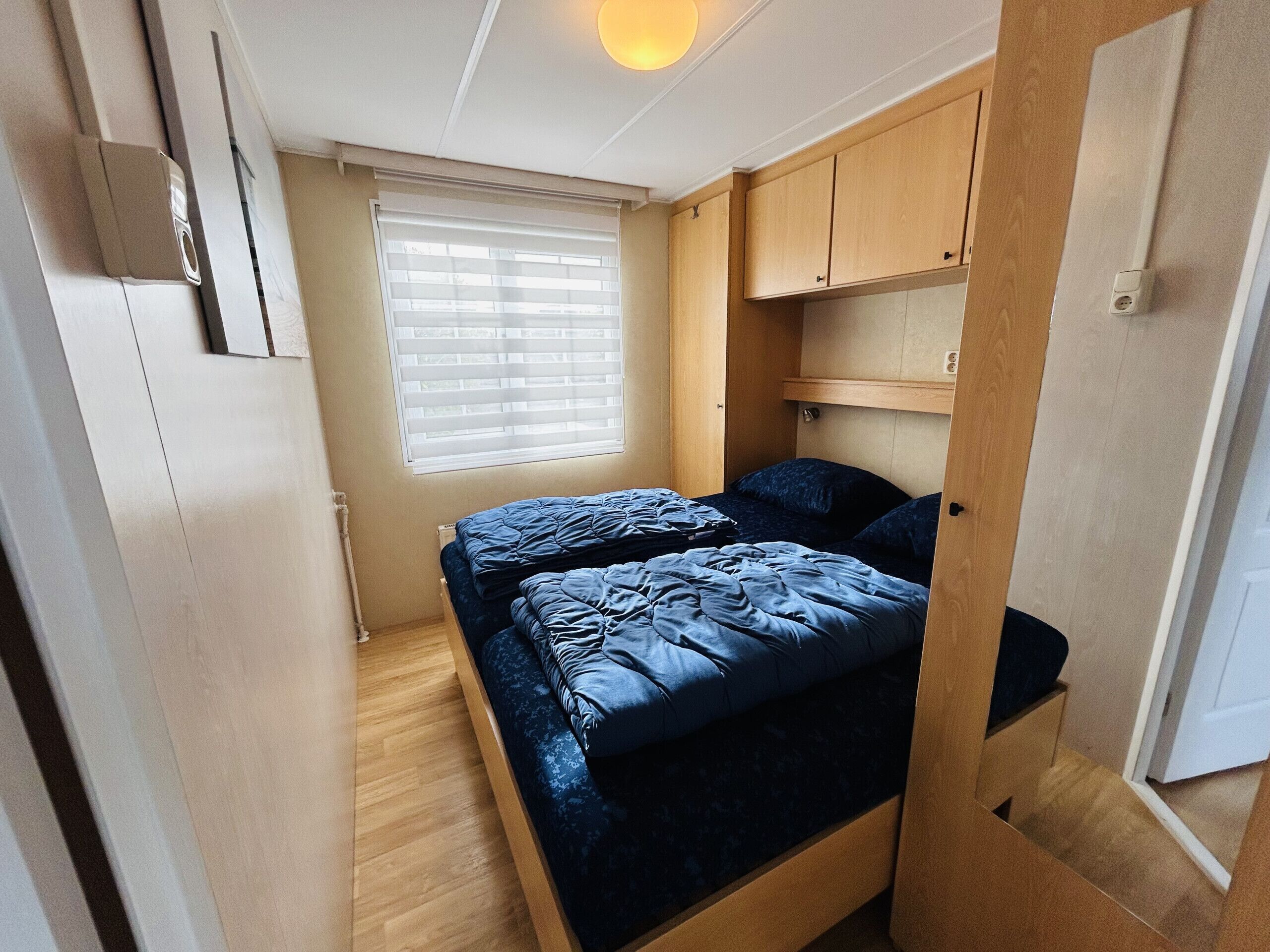 Quarto compacto com cama, janela com persianas e mobiliário de madeira.