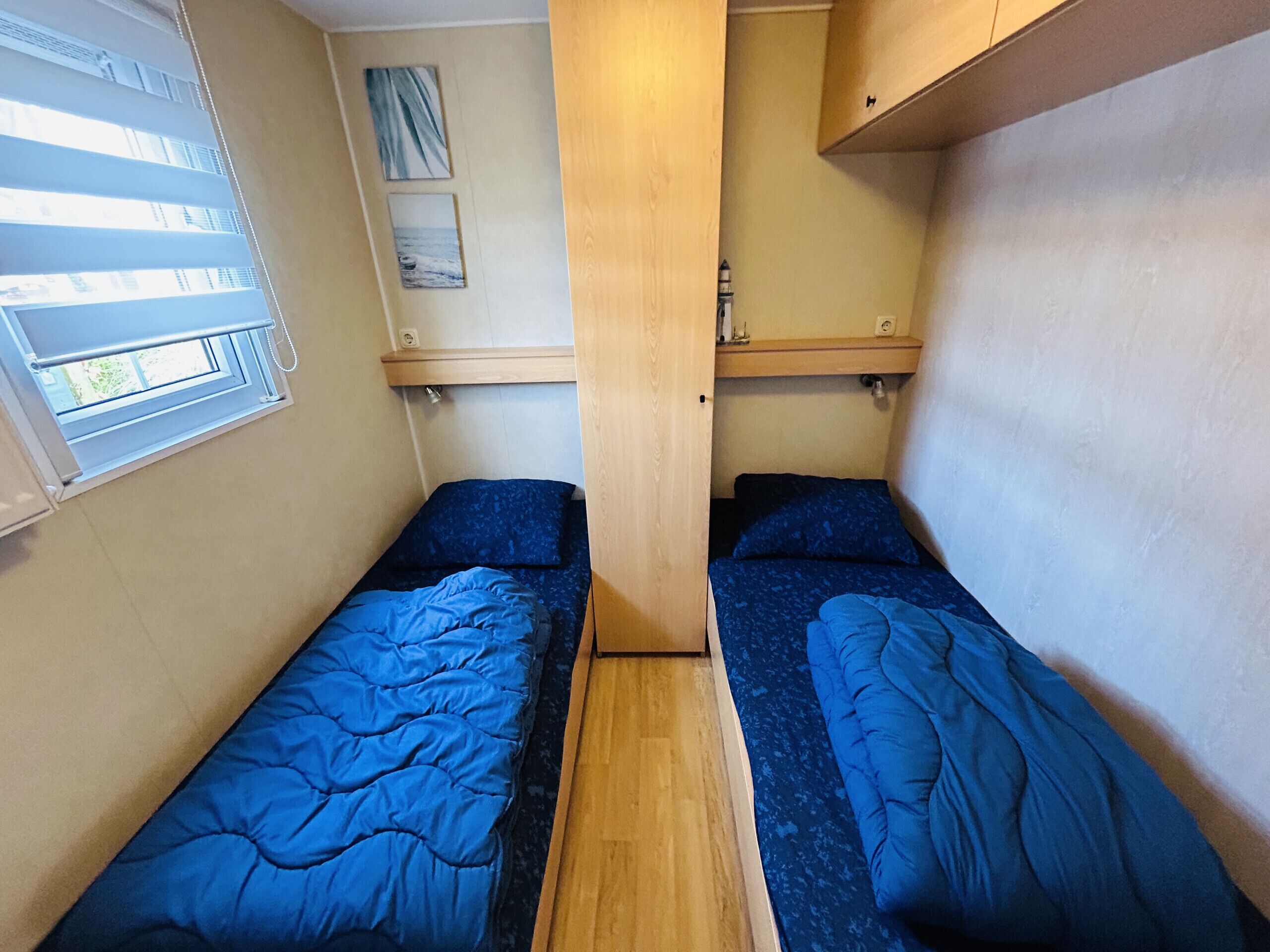 Una stanza compatta con due letti singoli, una piccola finestra e uno scaffale in legno.