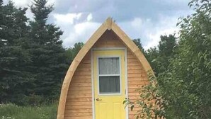 Una cabaña pequeña de madera con puerta amarilla y techo triangular, rodeada de follaje.