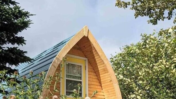 Una cabaña pequeña de madera con techo triangular, rodeada de abundante vegetación y plantas en flor.