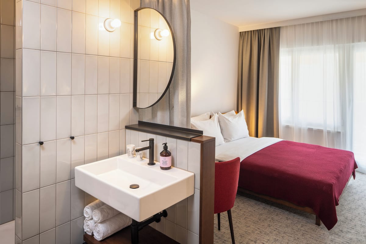 A hotel room with a large bed, a red chair, a white sink, and a mirror.