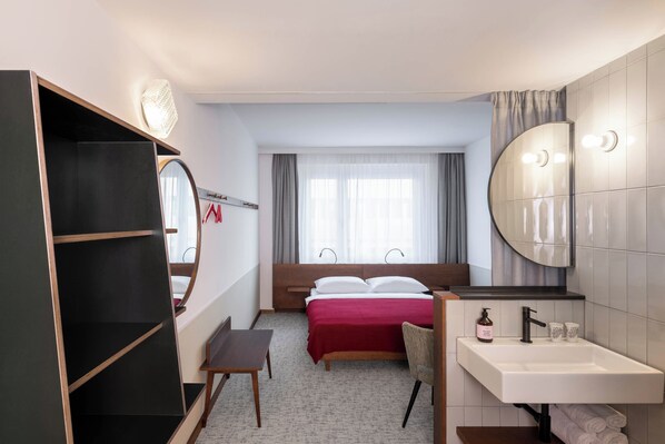 A hotel room with a bed, a desk, a wardrobe, a bathroom with a sink and mirror, and a window with curtains. - MAGDAS Hotel Vienna City (Vienna)