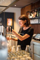 Um bartender prepara bebidas atrás do balcão, com um painel de madeira e prateleiras cheias de garrafas.