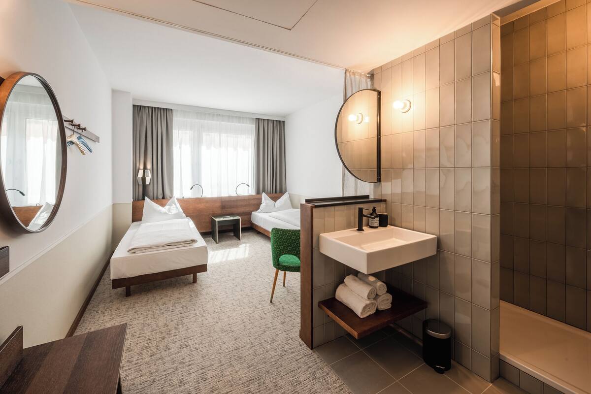 A hotel room with a bed, a small desk, a chair, a bathroom with a sink and mirror, and a shower area.