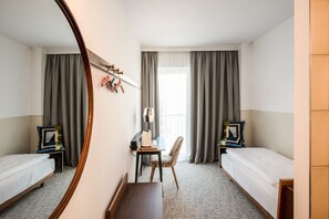 A hotel room with two single beds, a desk with a chair, a mirror, and a window with curtains.