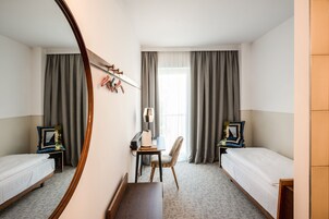 Quarto de hotel com duas camas de solteiro, uma escrivaninha com cadeira, um espelho e uma janela com cortinas.