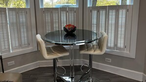 Une pièce d’angle avec une table en verre et deux chaises, un bol rempli d’en-cas et des volets blancs aux fenêtres.