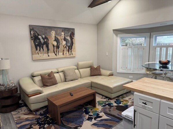 A living room with a sectional sofa, a coffee table, a rug, a painting of horses, and a view through a window.